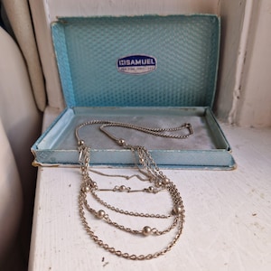 May include: A silver necklace with multiple strands and small beads, presented in an open, pale blue box. The box lid features the text "SAMUEL FOR FINE JEWELLERS".