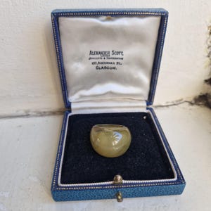 May include: A smooth, translucent, pale olive-green ring resting in a square blue velvet-lined presentation box. The interior lid features the printed text: Alexander Scott, Jewellers & Silversmiths, 109, Buchanan St., Glasgow.