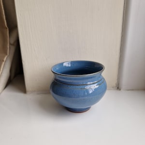 May include: A small, round ceramic bowl with a glossy, speckled blue glaze. The bowl features a flared rim, a curved body, and a narrow base. The interior and exterior surfaces show a consistent blue colour with subtle dark speckles throughout.