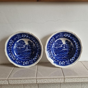 May include: Two blue and white ceramic plates featuring a landscape scene. The plates have a scalloped edge and a floral border. The central design shows a bridge, trees, and buildings. The plates are on a tiled surface.