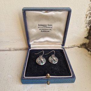 May include: A pair of silver-toned drop earrings featuring circular pendants with an iridescent abalone shell inlay and an openwork metal design. The earrings are presented in a blue hinged jewellery box with white satin lining and the text Alexander Scott Limited Jewellers & Silversmiths 109 Buchanan St. Glasgow.