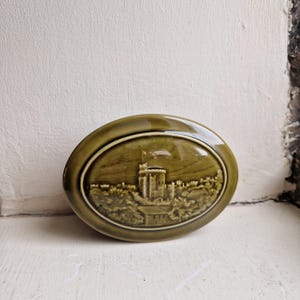 May include: An oval-shaped olive green ceramic trinket box featuring a raised relief design of a castle with a flag on the lid. The glossy finish highlights the detailed architectural structure of the fortress surrounded by trees and foliage.