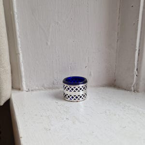 May include: A small, cylindrical container with a silver filigree design and a deep blue interior. The container is on a white windowsill. The design features circular cutouts. The container is likely for small items. The item is approximately 4 cm in height.
