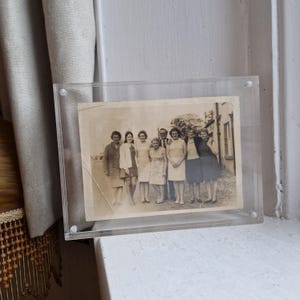May include: A vintage black and white photograph of a group of people, displayed in a clear acrylic frame. The framed photo is positioned on a white windowsill. The image shows a group of people, possibly family or friends, posing together.