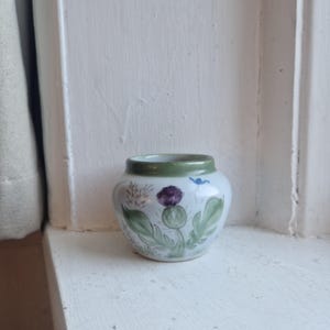 May include: A small, round ceramic vase with a white base and a green rim. The vase is decorated with a purple thistle flower, green leaves, and small blue accents. The vase is sitting on a white windowsill.