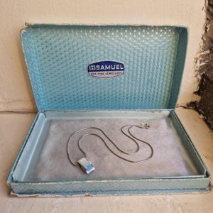 May include: A silver-toned pendant necklace with a rectangular metallic charm resting inside a light blue hinged jewellery box. The interior lid of the box features a geometric pattern and a blue label with the text H.SAMUEL FOR FINE JEWELLERY.