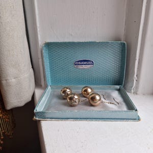 May include: A vintage jewellery box with a light blue interior contains two pairs of earrings. One pair has gold spheres, the other a soccer ball design. The box is labelled "SAMUEL FOR FINE JEWELLERS".