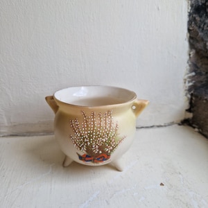May include: A small, round ceramic planter with a cream interior and a light yellow exterior. The planter features a floral design with white and green accents, a red and blue ribbon, and three small feet. It has two small handles.