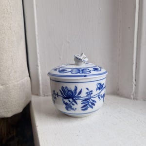 May include: A white ceramic sugar bowl with a blue floral pattern and a matching lid. The lid has a small, white, rose-shaped handle. The bowl and lid feature a detailed blue floral design.