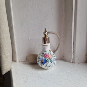 May include: A vintage white porcelain perfume bottle with a floral design in pink, blue, and yellow. The bottle has a gold-coloured metal top and a beige atomizer bulb. The bottle is approximately 13 cm tall.