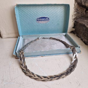 May include: A braided multi-tone metal necklace featuring interwoven silver and dark grey strands. The necklace is presented in a light blue rectangular jewellery box with a hinged lid, featuring the text H. SAMUEL FOR FINE JEWELLERS inside.