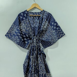 May include: A navy blue kaftan with a white patterned design, hanging on a wooden hanger. The kaftan features a V-neck and a tie closure at the waist. The fabric has a mix of geometric and floral patterns.