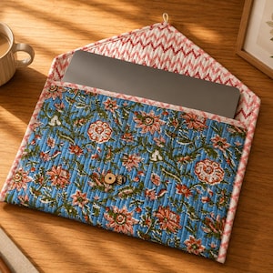 May include: A blue floral quilted laptop sleeve with a wooden button closure. The sleeve has a pink and white zigzag patterned lining. A cup of coffee sits to the left of the sleeve on a wooden surface.
