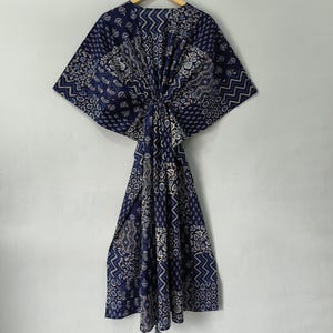 May include: A navy blue patterned kaftan dress with a geometric and floral design. The dress is hanging on a wooden hanger against a white wall. The kaftan features a loose, flowing silhouette.