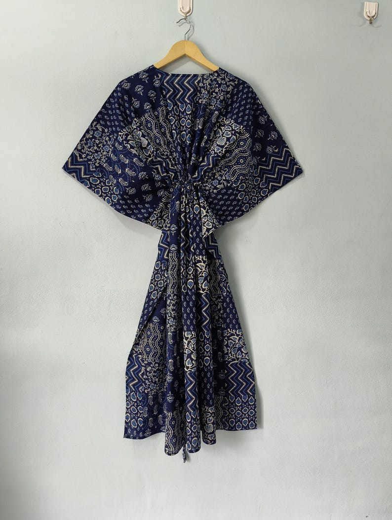 May include: A navy blue patterned kaftan dress with a geometric and floral design. The dress is hanging on a wooden hanger against a white wall. The kaftan features a loose, flowing silhouette.