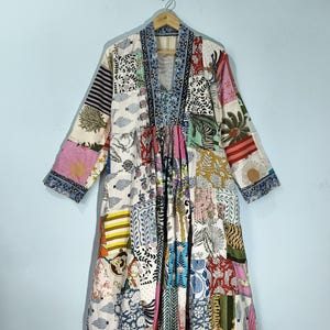 May include: A patchwork robe with a V-neck and long sleeves, hanging on a wooden hanger. The robe features a variety of colourful fabric patches with floral, animal, and geometric patterns. The sleeves and neckline are trimmed with a blue patterned fabric.