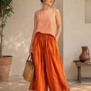 May include: A sleeveless orange and white striped top paired with wide-leg orange pants. The outfit is complemented by a woven tote bag and brown sandals. The ensemble suggests a casual, summery style.