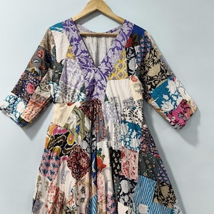 May include: A patchwork dress with a V-neck and three-quarter sleeves. The dress features a variety of floral and geometric patterns in a mix of colors including purple, blue, red, and white. The dress is hanging on a wooden hanger.