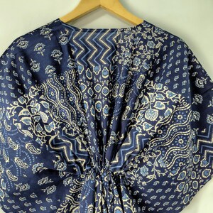 May include: A navy blue kaftan with a floral and geometric pattern in light blue and white. The fabric is gathered at the waist and hangs from a wooden hanger. The design includes paisley, floral, and zigzag patterns.