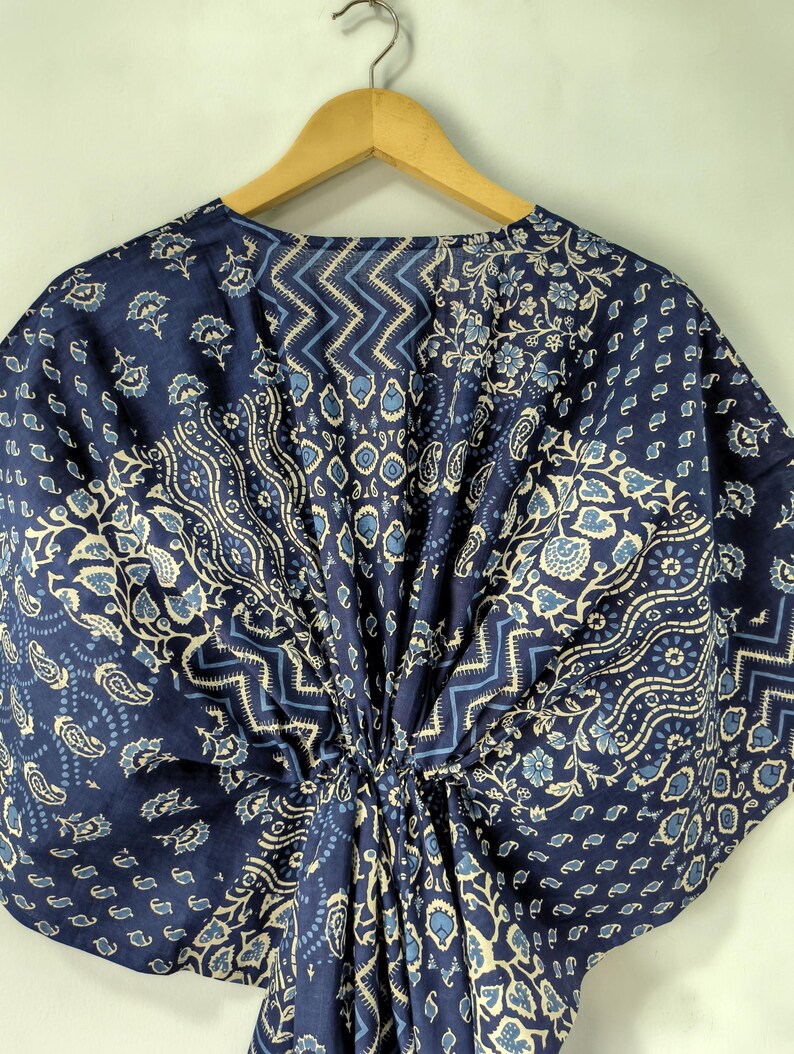 May include: A navy blue kaftan with a floral and geometric pattern in light blue and white. The fabric is gathered at the waist and hangs from a wooden hanger. The design includes paisley, floral, and zigzag patterns.