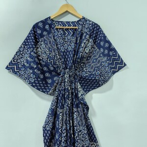 May include: A navy blue kaftan with a white and light blue patterned design. The kaftan is hanging on a wooden hanger against a light blue wall. The fabric has a paisley and geometric print.