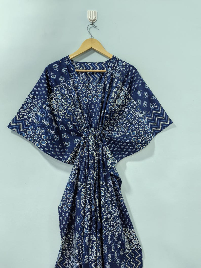 May include: A navy blue kaftan with a white and light blue patterned design. The kaftan is hanging on a wooden hanger against a light blue wall. The fabric has a paisley and geometric print.