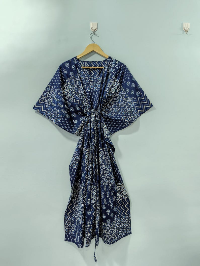 May include: A navy blue kaftan with a white patterned design, hanging on a wooden hanger. The kaftan features a V-neck and a tie closure at the waist. The fabric has a mix of geometric and floral patterns.