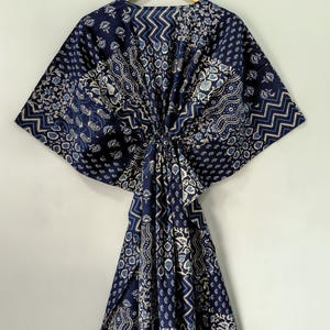 May include: A navy blue kaftan with a variety of white and light blue patterns, including floral, geometric, and paisley designs. The kaftan is hanging on a wooden hanger against a plain white background. The fabric is gathered at the waist.