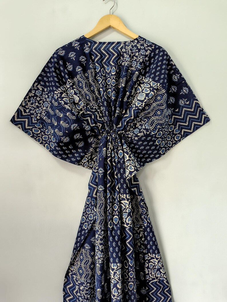 May include: A navy blue kaftan with a variety of white and light blue patterns, including floral, geometric, and paisley designs. The kaftan is hanging on a wooden hanger against a plain white background. The fabric is gathered at the waist.