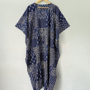 May include: A blue and white patterned caftan dress with short sleeves, hanging on a wooden hanger. The dress features a patchwork design with various geometric and floral patterns. The fabric appears lightweight and suitable for warm weather.