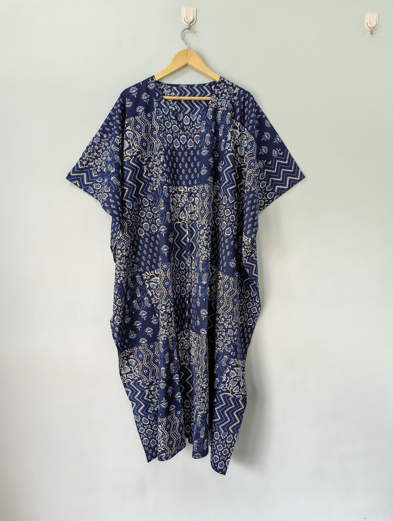 May include: A blue and white patterned caftan dress with short sleeves, hanging on a wooden hanger. The dress features a patchwork design with various geometric and floral patterns. The fabric appears lightweight and suitable for warm weather.