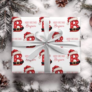 May include: White wrapping paper featuring a repeating pattern of the letter "B" in a Santa Claus hat and belt design. The words "Merry Christmas Bryson" are printed on the paper. A silver ribbon is tied around the wrapped gift.