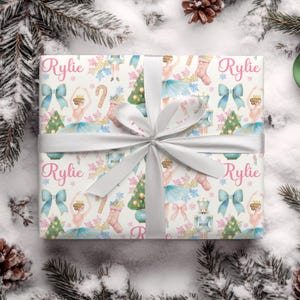 May include: A wrapped gift with a white ribbon and bow. The wrapping paper features a pattern of ballerinas, Christmas trees, bows, and snowflakes in pastel colors. The name "Rylie" is printed in pink on the wrapping paper.