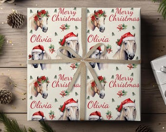 Puede incluir: Papel de regalo navideño con un patrón repetido de caballos con gorros de Papá Noel y coronas florales. Las palabras "Merry Christmas" y el nombre "Olivia" también están impresos. Una cinta dorada está atada alrededor del regalo.