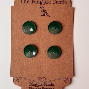 May include: Four round, dark green buttons with subtle glitter accents are attached to a rectangular brown cardboard display card. The card features the text The Magpie Curio at the top, a small bird illustration, and Magpie Finds Vintage Buttons at the bottom.