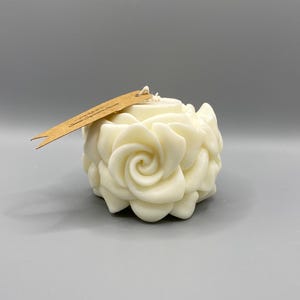 Può includere: Una candela bianca a forma di rosa con un'etichetta di carta marrone che dice "Glimmer Signature Scents".