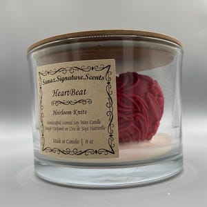 Può includere: Un barattolo di candela di vetro trasparente con coperchio in legno. All'interno, una candela rossa a forma di rosa poggia su una base bianca. L'etichetta recita "HeartBeat" e "Heirloom Knits". La candela è di 227 grammi.