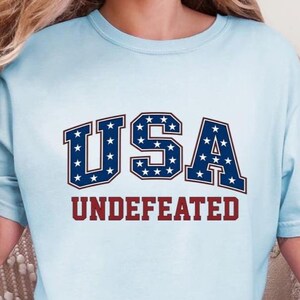 May include: Light blue t-shirt with the word "USA" in navy blue block letters with white stars, and the word "UNDEFEATED" in red block letters. The shirt is a casual style.