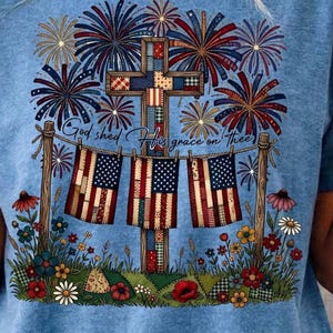 Può includere: T-shirt azzurra con un design patriottico. Il design include una croce, bandiere americane, fuochi d'artificio ed elementi floreali. È presente anche il testo "God shed His grace on thee".