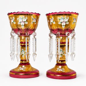 May include: Two antique, ornate chalices with gold-colored bowls and ruby red rims and bases. The bowls are decorated with hand-painted white and blue flowers. Clear crystal prisms hang from the bowls. The chalices are on a white background.