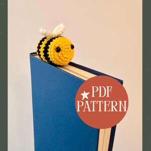 May include: A handmade crochet bee bookmark in yellow and black, with white wings, rests on a blue book. The bee is positioned at the top of the book, with a round orange sticker that says "PDF PATTERN".