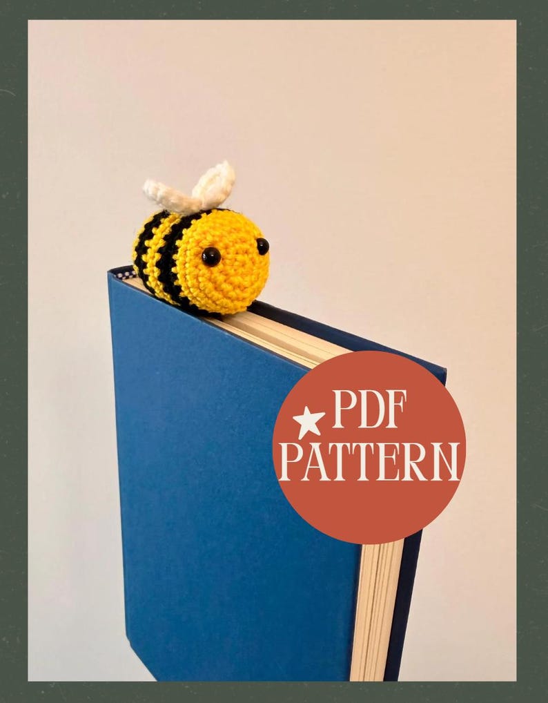 May include: A handmade crochet bee bookmark in yellow and black, with white wings, rests on a blue book. The bee is positioned at the top of the book, with a round orange sticker that says "PDF PATTERN".