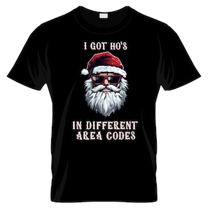 May include: Black t-shirt featuring a Santa Claus graphic wearing sunglasses and a red hat. The text above the graphic reads "I GOT HO'S" and below reads "IN DIFFERENT AREA CODES".