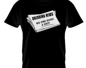 May include: Black t-shirt featuring a graphic of a newspaper with the headline "BREAKING NEWS" and the text "NO ONE GIVES A SHIT!". The design is in black and white, contrasting against the black fabric of the shirt.