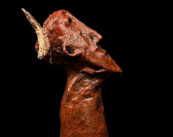 Könnte beinhalten: Eine rote und goldene Skulptur eines menschlichen Kopfes mit einem einzigen Horn. Die Skulptur ist aus einem rauen, strukturierten Material gefertigt und hat einen verwitterten Look.