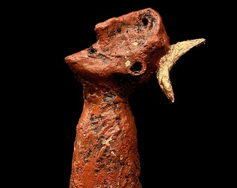 Könnte beinhalten: Eine rote und goldene Keramikskulptur eines stilisierten menschlichen Kopfes mit einem einzigen Horn. Die Skulptur hat eine raue, strukturierte Oberfläche und ist in Rot- und Goldtönen bemalt.