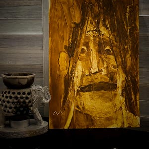 May include: Abstract oil painting on canvas, featuring a face with a neutral expression. The artwork uses shades of brown and gold, with visible brushstrokes. A small, carved elephant statue and a decorative bowl are in the foreground.