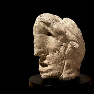 May include: A sculpted bust of a human head in profile, crafted from a light-colored material. The sculpture has a rough, textured surface and sits on a small, round black base against a dark background. The artwork captures the likeness of a person.