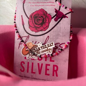 May include: A beaded necklace in pink and black with a charm reading "Wear the hat, Ride the cowboy" lies on a book cover featuring a rose illustration. The book is titled "Royal" by Elsie Silver.
