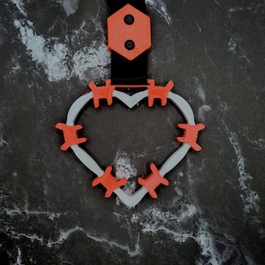 Puede incluir: Un llavero en forma de corazón impreso en 3D con detalles en naranja. El corazón está delineado en gris y presenta adornos en forma de gato naranja alrededor del perímetro. El llavero tiene una correa negra y una pieza superior hexagonal naranja con dos círculos negros.